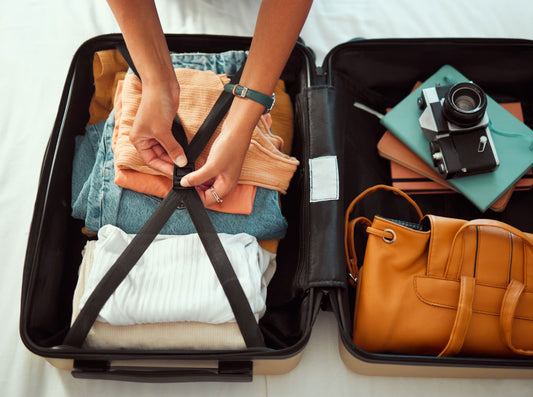Hands pressing folded clothes into an open suitcase under its cross straps, with a camera, notebook and tan handbag in the other half