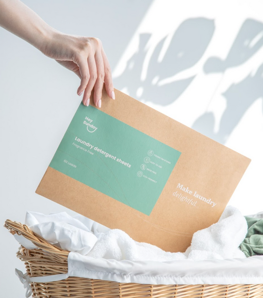 Hand holding a HeySunday Fragrance Free laundry detergent sheets envelope above a wicker basket of white laundry