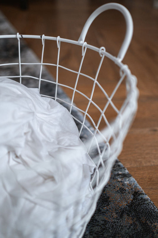 A white wire laundry basket with crumpled clothes placed on a hardwood floor inside a home