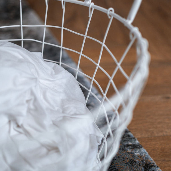 A white wire laundry basket with crumpled clothes placed on a hardwood floor inside a home