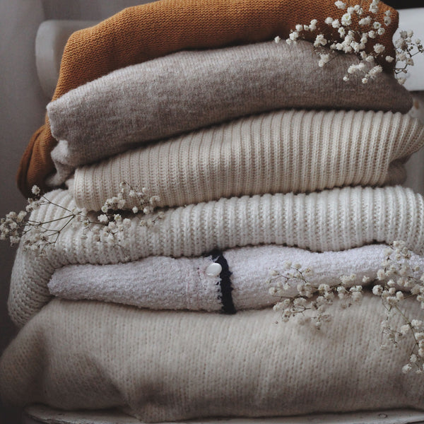 Aesthetic stack of beige and brown woolen sweaters on a chair indoors, adorned with dried flowers.